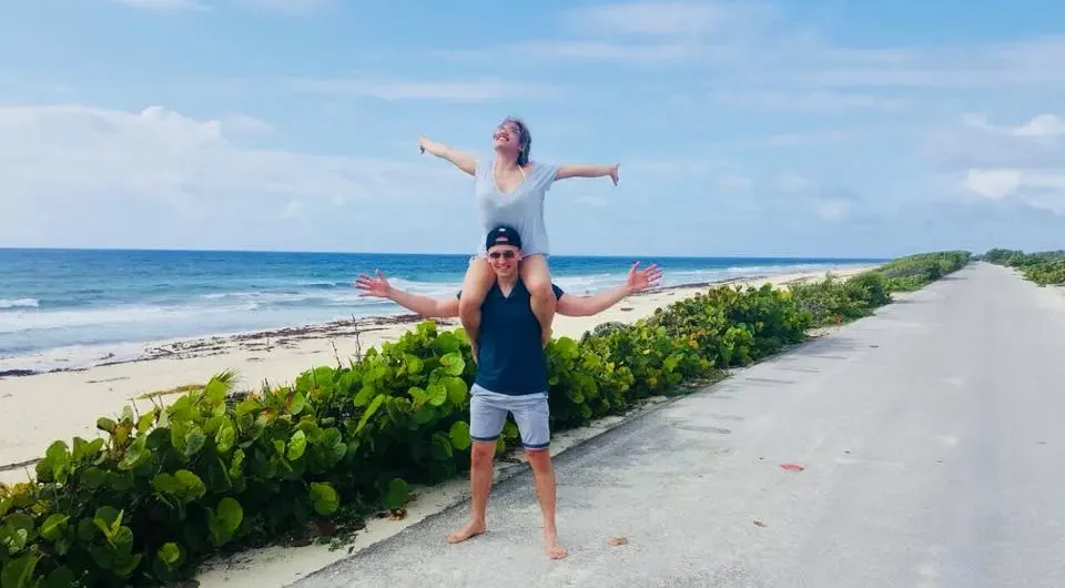 A happy young couple laughing on the scenic coastal road of Cozumel's East Side, with the woman on the man's shoulders, during a fun private jeep tour.