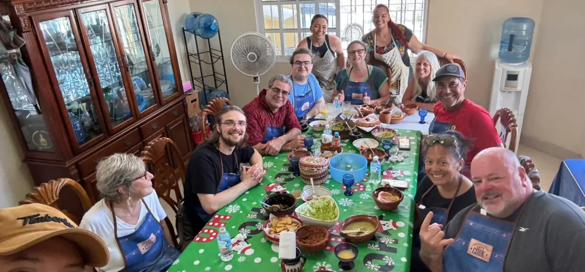A diverse group of happy travelers enjoying a meal they prepared together around a festive table during an authentic market-to-table cooking class in a local Cozumel home.