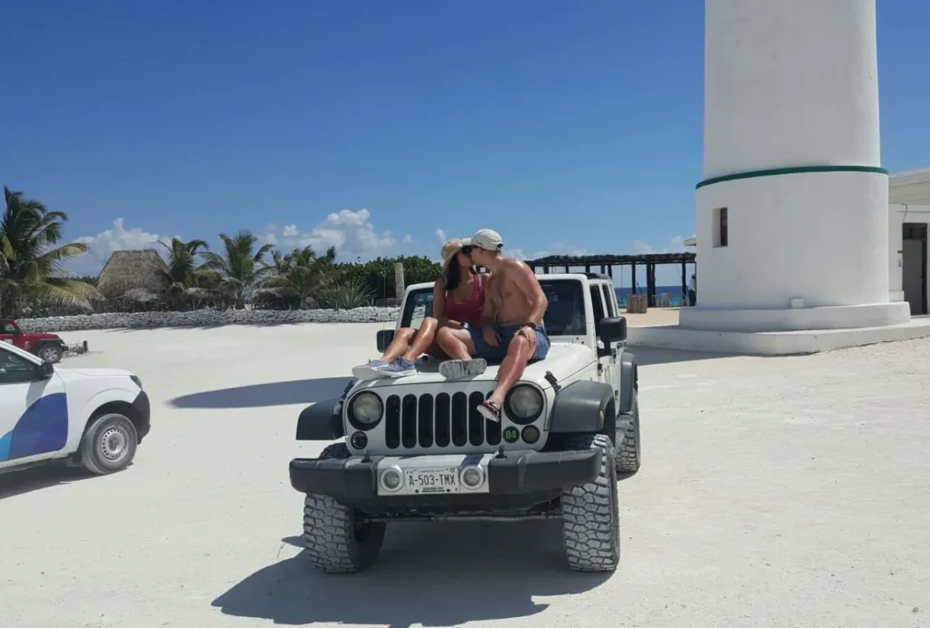 A romantic couple kissing on the hood of a white Jeep Wrangler in front of the iconic Celarain lighthouse at Punta Sur Eco Beach Park, Cozumel, on a sunny day.