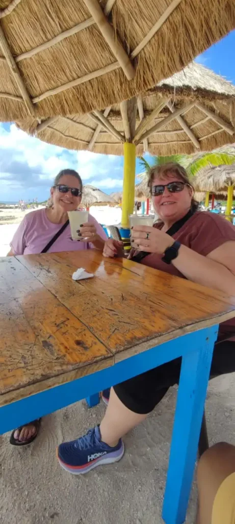 Two happy women enjoying refreshing drinks under a palapa at a rustic beach bar in Cozumel, relaxing after a family-friendly jungle adventure tour.