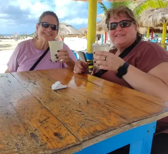 Two happy women enjoying refreshing drinks under a palapa at a rustic beach bar in Cozumel, relaxing after a family-friendly jungle adventure tour.