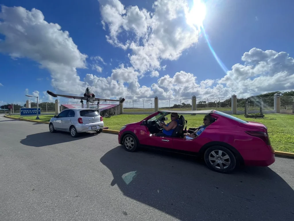 Two guests enjoying a sunny day on their Cozumel excursion, driving a bright pink convertible buggy near the air force monument.
